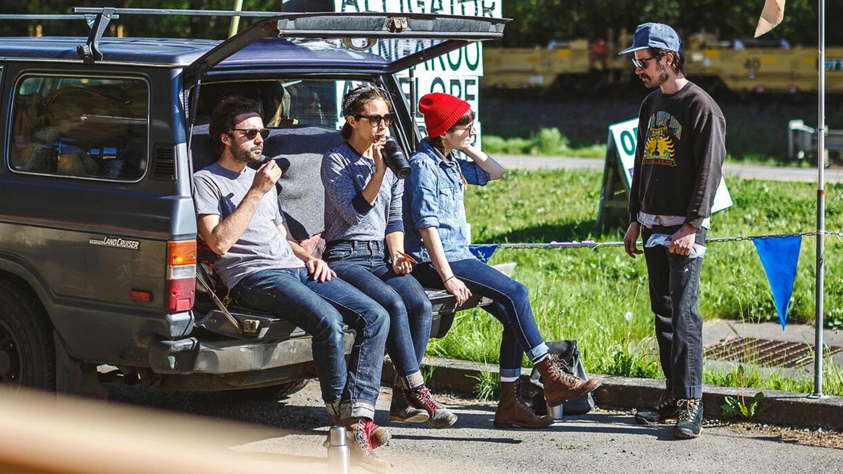 Three hikers hanging out in the tailgate of a station wagon while a fourth hiker looks on.